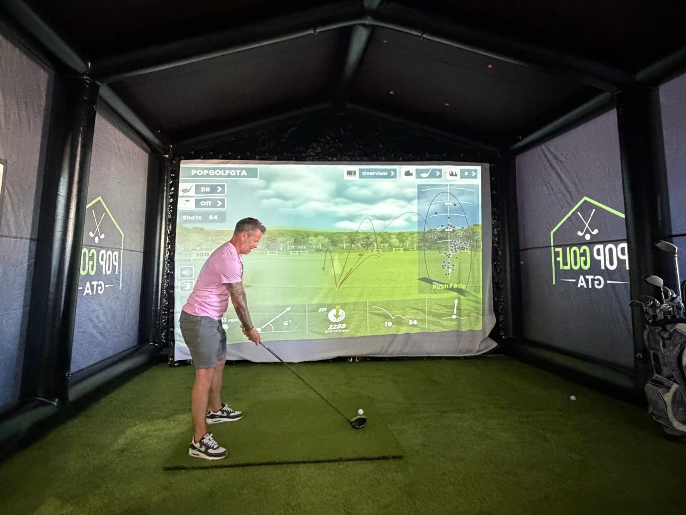 Guest teeing off inside the Pop Golf GTA simulator enclosure, with ball-flight tracking on the big screen